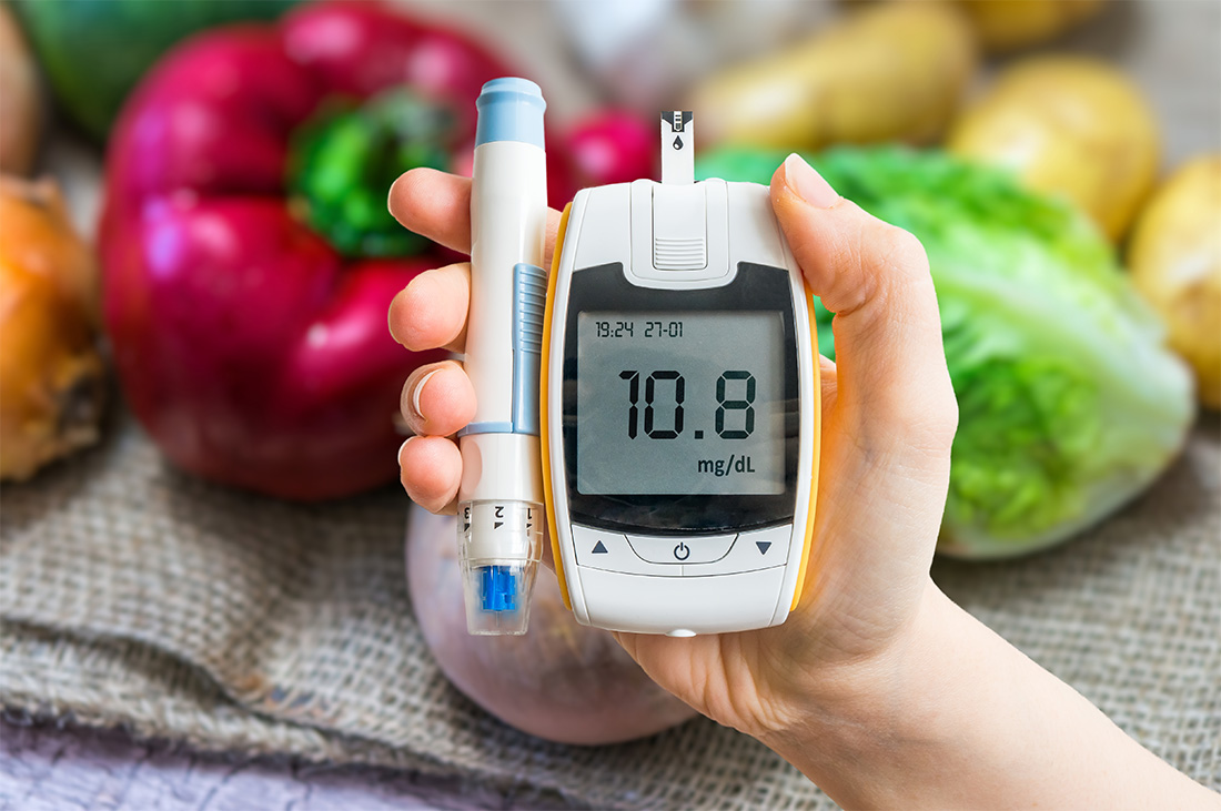 Hand holding glucometer and pen with vegetables in background.